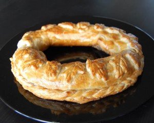 A freshly baked, golden brown pretzel-shaped pastry on a black plate, with a flaky texture visible on its surface, reflecting light.