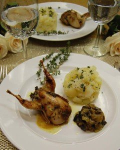 A gourmet plate featuring roasted quail, mashed potatoes, and a mushroom side dish, elegantly presented on a white plate with a glass of water, wine, and roses on the table.
