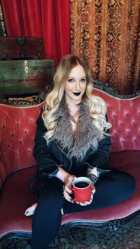 A woman with long blonde hair, wearing a black coat and gray scarf, sits on a red tufted sofa holding a red mug, against a backdrop of richly patterned curtains.