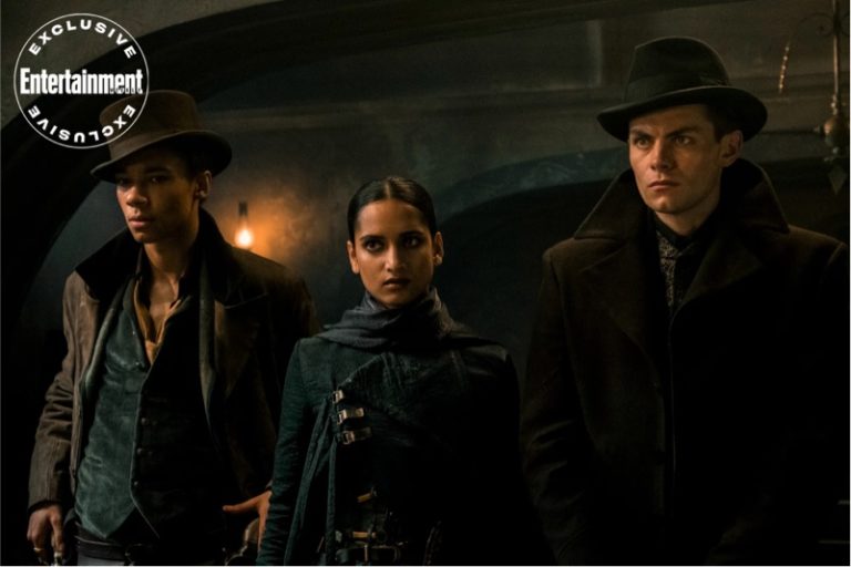 Three actors in period costumes stand in a dimly lit, atmospheric setting, the center one a woman flanked by two men, all conveying a serious demeanor. a logo reads "entertainment exclusive.